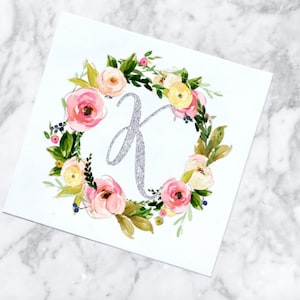 May include: A white card with a floral wreath surrounding a silver glitter letter K. The wreath is made of pink, white, and yellow flowers with green leaves.