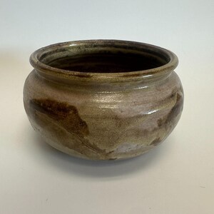 Puede incluir: Un cuenco de cerámica hecho a mano con forma redondeada y una amplia abertura. El cuenco presenta una mezcla de tonos terrosos, incluyendo marrón, beige y toques de rosa. El exterior tiene una apariencia texturizada, y el borde es marrón oscuro.