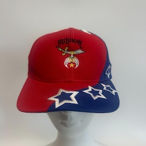 May include: Red and blue baseball cap with embroidered text "Medinah" and a symbol featuring a sword, crescent moon, and star. The brim has white embroidered stars on a blue background.