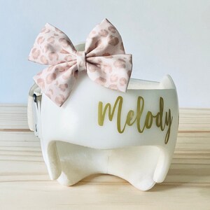 May include: White helmet with a pink and brown leopard print bow on top. The helmet has the name "Melody" written in gold letters on the front.