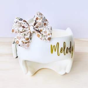 May include: White padded headband with a floral print bow and the name "Melody" written in gold lettering.