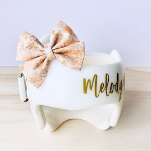 May include: White padded helmet with a gold vinyl name "Melody" on the side. A floral patterned bow is attached to the top of the helmet.