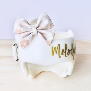 May include: White padded headband with the name "Melody" written in gold lettering. A pink and white floral bow is attached to the headband.
