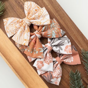 May include: Three fabric hair bows with floral patterns in shades of orange, white, and green. The bows are arranged on a wooden surface.