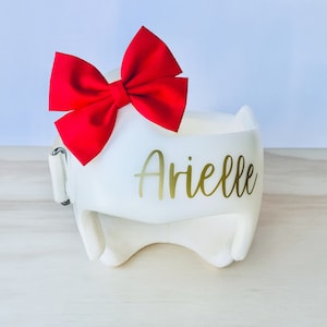 May include: White padded headband with a red bow and the name "Arielle" written in gold lettering.