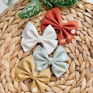 May include: Four fabric hair bows with white flowers and gold lettering. The bows are in the colors white, light green, mustard yellow, and burnt orange. The bows are on a woven wicker surface. The bows are personalized with the names "Lainey", "Riley", and two other names.