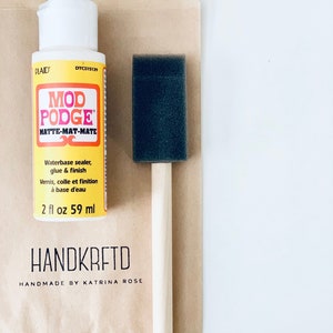 May include: A white bottle of Mod Podge Matte-Mat-Mate glue and sealer with a black foam brush. The bottle is labeled with the brand name "Plaid" and the product name "Mod Podge". The bottle contains 2 fluid ounces (59 ml) of product. The label also includes the text "Waterbase sealer, glue & finish" and "Vernis, colle et finition à base d'eau". The bottle is sitting on a brown paper bag with the text "HANDKRETD" and "HANDMADE BY KATRINA ROSE" printed on it.