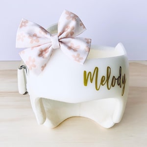 May include: White helmet with a pink floral bow and the name "Melody" written in gold lettering.