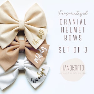 May include: Set of three personalized cranial helmet bows in beige, brown, and white with gold lettering. The bows are labeled "Charlotte", "Melody", and "Evelyn".