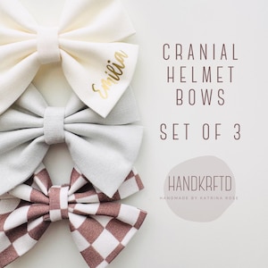 May include: Set of three cranial helmet bows. One bow is white with the name "Emilia" written in gold lettering. Another bow is light gray. The third bow is a brown and white checkered pattern.