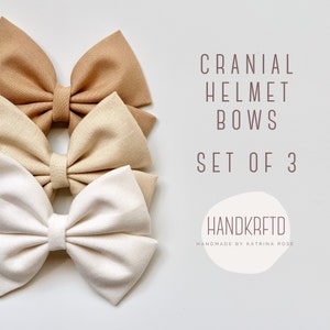 May include: Set of three fabric bows in beige, light brown, and white. The bows are arranged in a stack, with the largest bow on the bottom and the smallest bow on top. The text "Cranial Helmet Bows Set of 3" is written in brown text above the bows. The text "HANDKRFETD Handmade by Katrina Rose" is written in brown text below the bows.