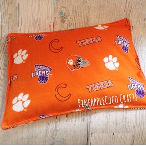 May include: Orange and white fabric pillow with a Clemson Tigers design. The pillow has a paw print, a tiger head, and the words "Tigers" and "C" printed on it. The pillow is made by Pineapplecoco Crafts.