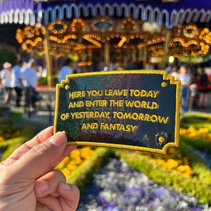 May include: A black rectangular patch with gold trim and the text "HERE YOU LEAVE TODAY AND ENTER THE WORLD OF YESTERDAY, TOMORROW AND FANTASY." The patch has a holographic effect. The background shows a carousel and flowers.