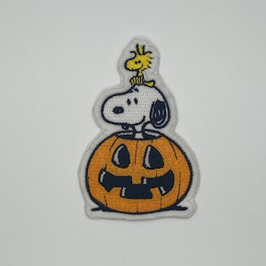 May include: An embroidered patch featuring Snoopy sitting on top of a jack-o'-lantern. Snoopy is wearing his signature red collar and is looking to the right. The jack-o'-lantern is orange with a black face and a wide grin.