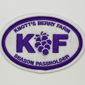 May include: White oval patch with purple stitching. The patch says "Knott's Berry Farm" and "Season Passholder" in a purple font. The letters "K" and "F" are large and there is a purple blackberry graphic between them.