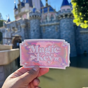 May include: A pink iridescent patch with white embroidery that reads "Magic Key" in a retro font. The patch is held in front of a blurred image of Sleeping Beauty's castle.