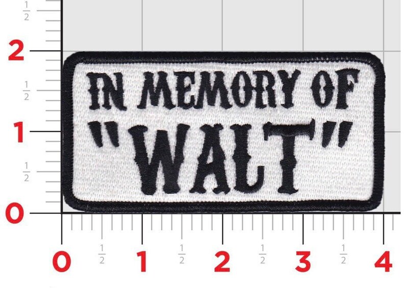 Op de afbeelding: Witte rechthoekige patch met zwarte geborduurde tekst die "In Memory Of "Walt" luidt.