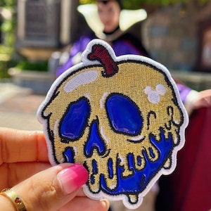 May include: A yellow and blue embroidered patch shaped like a poisoned apple with a skull and crossbones design. The apple is dripping blue liquid.