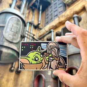 May include: A black and white embroidered patch featuring a green alien character with large eyes and a brown Mandalorian character with a helmet. The patch has a blue and white background with red and green lights in the top left corner.