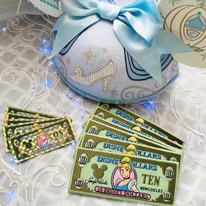 May include: Six green and gold embroidered patches with Cinderella and Mickey Mouse. The patches are shaped like dollar bills and say "10 Disney Dollars" and "Ten".