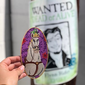 May include: An oval embroidered patch featuring a white horse with a stern expression and a small green lizard on its head, set against a purple background with red floral accents and a gold border, held up against a blurred wanted poster.
