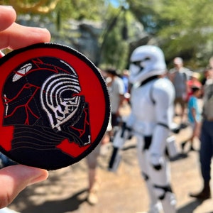May include: A red and black embroidered patch featuring a silhouette of Kylo Ren's helmet from Star Wars. The helmet is black with red accents and a white design on the front.