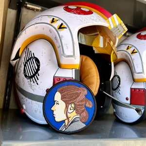 May include: A white and red pilot helmet with a yellow stripe and a blue and black embroidered patch of a woman's face. The patch is circular and has a blue background.