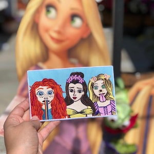 May include: An embroidered patch featuring three Disney princesses:  Ariel, Belle, and Rapunzel.  Ariel is drinking from a rainbow straw, Belle is smiling, and Rapunzel has a dog nose and tongue sticking out.