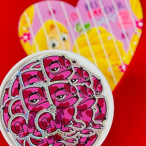 May include: A stack of pink and white glitter stickers featuring a cartoon chameleon with a heart pattern. The chameleon has a grumpy expression and is looking to the left.