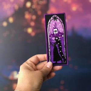 May include: A rectangular embroidered patch featuring a purple and black design of a silhouette with long hair standing before an arched window. The scene depicts a castle tower under a night sky with a sun emblem at the top and stars throughout.