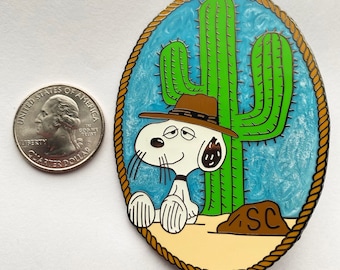 Op de afbeelding: Een cartoonhond met een cowboyhoed zit naast een groene cactus in een woestijnlandschap. De hond is wit met zwarte ogen en een zwarte neus. De cactus is hoog en heeft een bruine basis. De achtergrond is blauw en wit. De tekst "SC" staat op de grond.