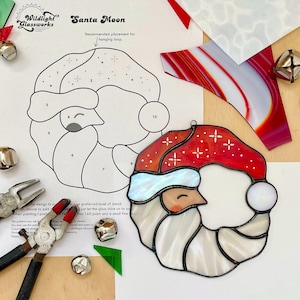 Puede incluir: Un adorno de Santa Moon de vidrieras con un gorro rojo y barba blanca. El diseño incluye un patrón y herramientas para crear el adorno. El texto "Santa Moon" y "Wildlight Glassworks" son visibles.