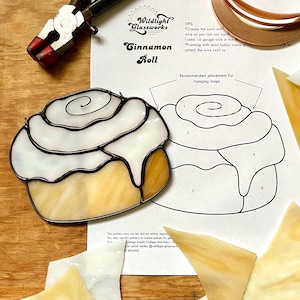 Puede incluir: Un patrón de rollo de canela de vitral con seis piezas de vidrio y un contorno negro. El patrón está en una hoja de papel blanca con instrucciones para los bucles colgantes y consejos para crear el proyecto de vitral. El texto en el papel dice "Wildlight Glassworks" y "Cinnamon Roll".