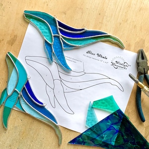May include: Two stained glass blue whale suncatchers with shades of teal, turquoise, and dark blue glass joined by silver solder. A paper pattern labelled Blue Whale measuring 25.4 cm by 11.4 cm, glass cutting tools, and glass shards.