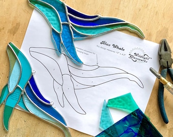 May include: Two stained glass blue whale suncatchers with shades of teal, turquoise, and dark blue glass joined by silver solder. A paper pattern labeled Blue Whale measuring 10 inches by 4.5 inches, glass cutting tools, and glass shards.