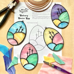 May include: A stained glass Easter egg craft kit with a pattern of three crosses on a hill. The kit includes a template, glass pieces, lead came, and instructions. The egg is a light pink, yellow, and blue colour.