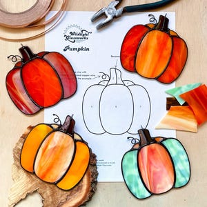 Pode incluir: Abóboras de vitral em tons de laranja, vermelho e verde, com contornos pretos. Elas estão dispostas em torno de um diagrama e ferramentas, sugerindo um projeto artesanal. O texto "Wildlight Glassworks Pumpkin" é visível.