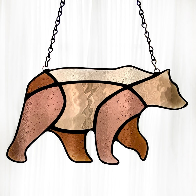 May include: A stained glass bear silhouette with brown and beige glass pieces. The bear is hanging from a black chain.