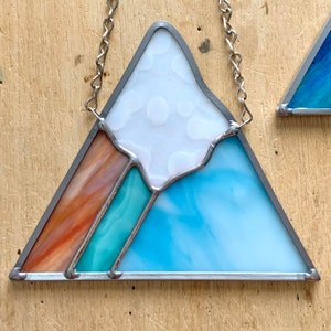 K&ouml;nnte beinhalten: Eine Berglandschaft aus Buntglas mit einem wei&szlig;en Gipfel, einem t&uuml;rkisfarbenen Sockel und einem braunen und orangen Streifen. Der Berg ist mit silbernem Metall umrahmt und hat eine silberne Kette, die an der Spitze befestigt ist.