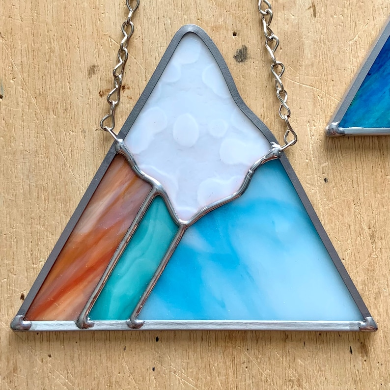 K&ouml;nnte beinhalten: Eine Berglandschaft aus Buntglas mit einem wei&szlig;en Gipfel, einem t&uuml;rkisfarbenen Sockel und einem braunen und orangen Streifen. Der Berg ist mit silbernem Metall umrahmt und hat eine silberne Kette, die an der Spitze befestigt ist.