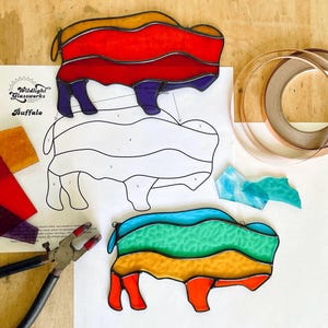 Pode incluir: Peças de arte de vitrais coloridos de búfalo em vermelho, laranja, verde e azul. A imagem inclui um padrão, peças de vidro e ferramentas para criar arte em vitrais. O logotipo Wildlight Glassworks é visível.