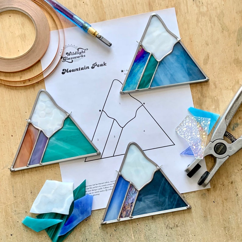 K&ouml;nnte beinhalten: Ein Stained-Glass-Bastelset f&uuml;r einen Berggipfel mit drei vorgeschnittenen Glasscheiben, einem Muster, Kupferfolie, einem Glasschneider und einer Zange. Das Set ist mit "Wildlight Glassworks Mountain Peak" beschriftet.