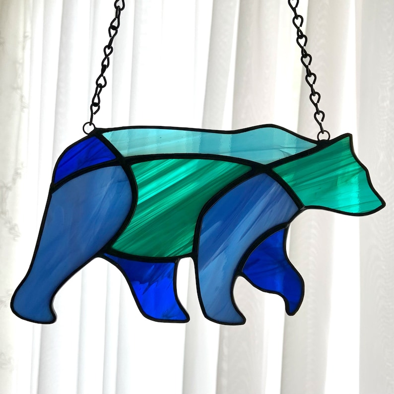 May include: A stained glass bear silhouette hanging from a chain. The bear is made of blue, green, and light blue glass.