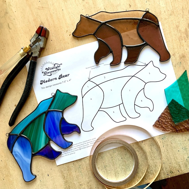 May include: A stained glass bear pattern with numbered sections for assembly. The pattern is printed on white paper with the text "Wildlight Glassworks Modern Bear This design measures 7.5" x 4" ". A brown stained glass bear, a blue and green stained glass bear, and a piece of copper tape are also shown.