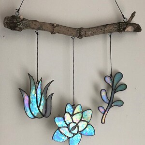 Op de afbeelding: Een mobiel van glas-in-lood met drie hangende delen. De mobiel is gemaakt van iriserend glas en heeft een succulent, een blad en een stengel. De mobiel hangt aan een natuurlijke tak.