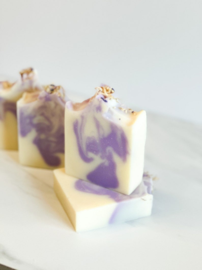 Op de afbeelding: Drie handgemaakte zeepjes met een swirl van paars en wit. De zeep is bedekt met gedroogde lavendelbloemen.