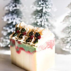 May include: A rectangular bar of handmade soap with a festive holiday design. The soap features layers of white, red, and green, topped with decorative cream-colored swirls and sprigs of holly with red berries. The background includes blurred, snow-covered Christmas trees.