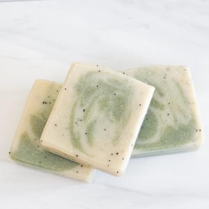 Op de afbeelding: Drie handgemaakte zeepjes met een groen-wit swirl-patroon. De zeep heeft een gespikkeld uiterlijk met kleine zwarte vlekjes.