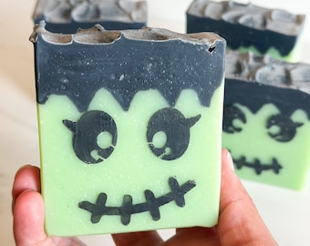 May include: Hand holding a green and black soap bar with a Frankenstein face design. The soap has a green base with a black top and a black face design. Other soap bars are in the background.