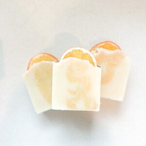 May include: Three white bars of soap with orange slices on top. The soap is a light white color with a subtle orange hue.